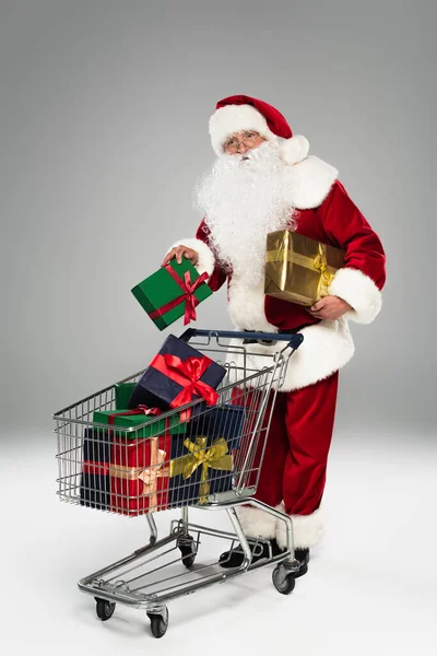Gri arka planda alışveriş arabasının yanında kostüm içinde tam boy Noel Baba. 