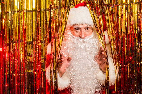 Санта Клаус в костюме смотрит в камеру возле блестящей мишуры 