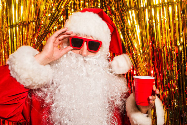 Санта-Клаус в солнцезащитных очках и шляпе с пластиковой чашкой возле мишуры 