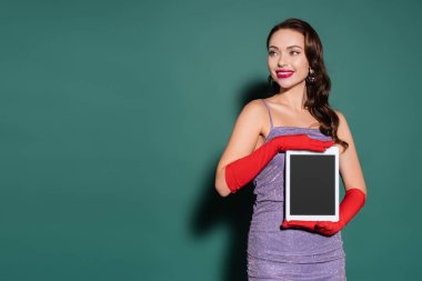 Mor elbiseli, eldivenli, elinde yeşil ekranlı dijital tablet olan neşeli genç bir kadın.