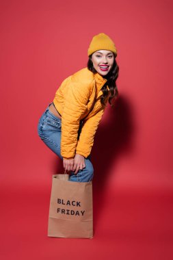 Kırmızı şapkalı ve kabarık ceketli mutlu genç bir kadın. Üstünde siyah cuma harfleri olan alışveriş çantasının içinde duruyor.