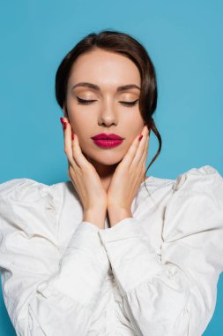 Beyaz bluzlu çekici genç bir kadının portresi. Gözleri kapalı poz veriyor ve mavi renkte izole edilmiş yüzlere dokunuyor.