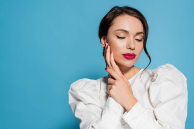 Beyaz bluzlu şehvetli genç bir kadının portresi. Kapalı gözlerle poz veriyor ve mavi üzerine izole edilmiş yüzlere dokunuyor.