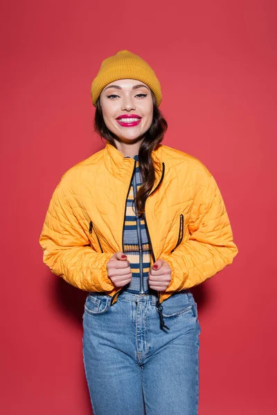 happy young woman in beanie hat adjusting puffer jacket and smiling on red