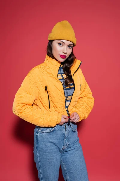 young woman in beanie hat zipping orange puffer jacket and looking at camera on red