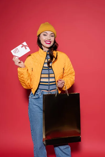 happy young woman in orange beanie hat holding gift card and shopping bag on red