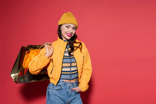 cheerful young woman in orange beanie hat and puffer jacket holding shopping bag on red