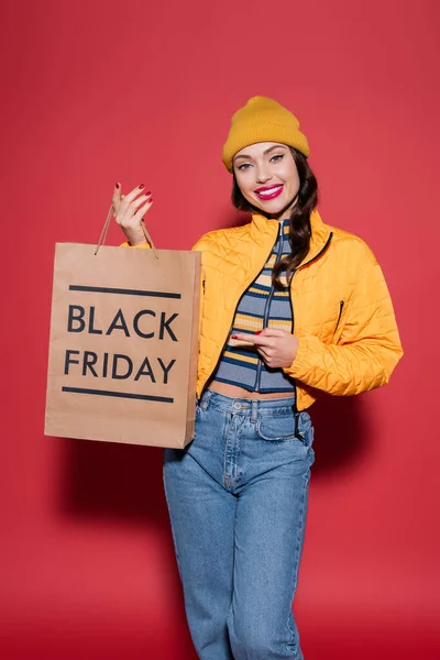 happy young woman in orange beanie hat and puffer jacket pointing at shopping bag with black friday lettering on red
