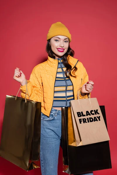 happy young woman in beanie hat and puffer jacket holding shopping bags with black friday lettering on red