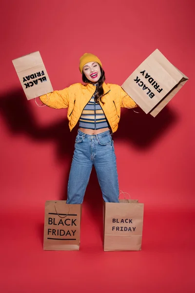 cheerful woman in beanie hat and puffer jacket with black friday shopping bags on hands standing on red