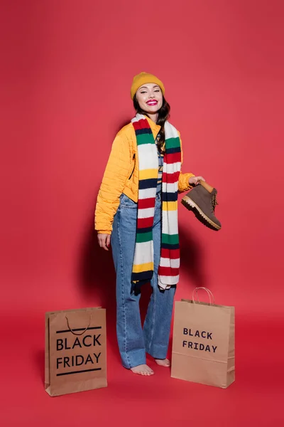 barefoot woman in puffer jacket standing with boot near shopping bags with black friday lettering on red