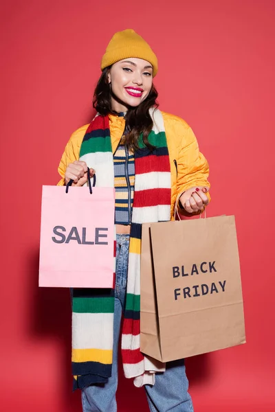 joyful woman in beanie hat and scarf holding shopping bags with black friday lettering on red
