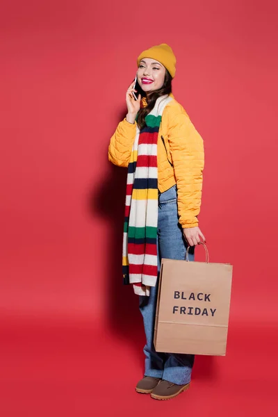 happy woman in beanie hat holding shopping bag with black friday lettering and talking on smartphone on red