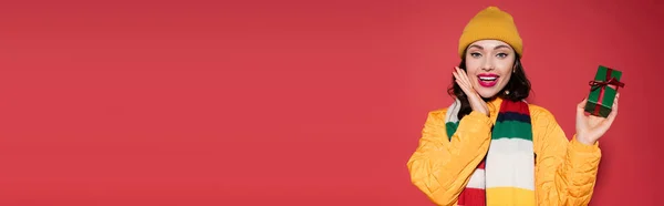 Изумленная Женщина Шапочке Шарфе Держащая Завернутый Подарок Красном Баннер — стоковое фото