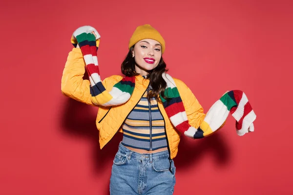 cheerful young woman in beanie hat and orange puffer jacket holding stripped scarf on red