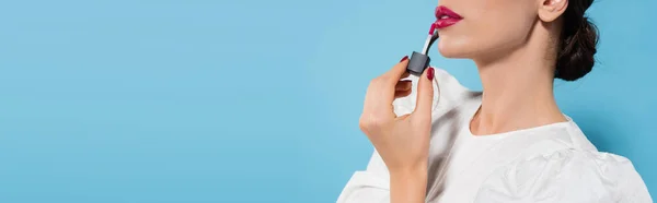 cropped view of young woman in blouse applying pink lip gloss isolated on blue, banner