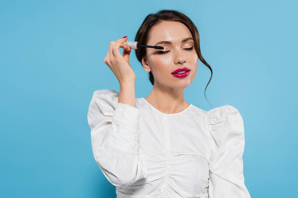 pretty young woman in white blouse applying mascara on eyelash on blue background