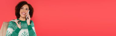 Sıcak kazak giyen neşeli Afro-Amerikalı kadın elinde çantalar ve kırmızı pankartta izole edilmiş akıllı telefondan konuşuyor.