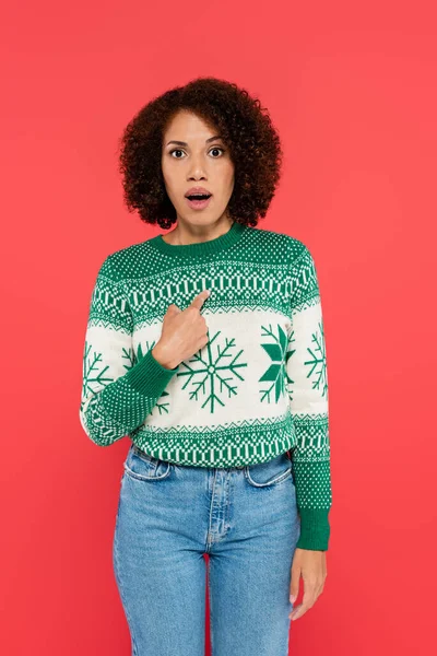 surprised african american woman in warm sweater with winter pattern pointing with finger at herself isolated on red