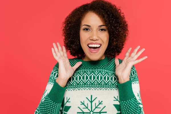 excited african american woman in winter sweater with green pattern showing wow gesture isolated on red