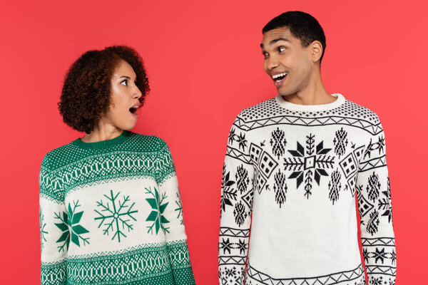astonished african american couple in warm sweaters looking at each other isolated on red