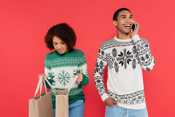 african american woman looking into shopping bags near cheerful man talking on cellphone isolated on red