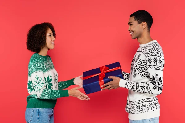 side view of african american man presenting christmas gift to happy girlfriend isolated on red