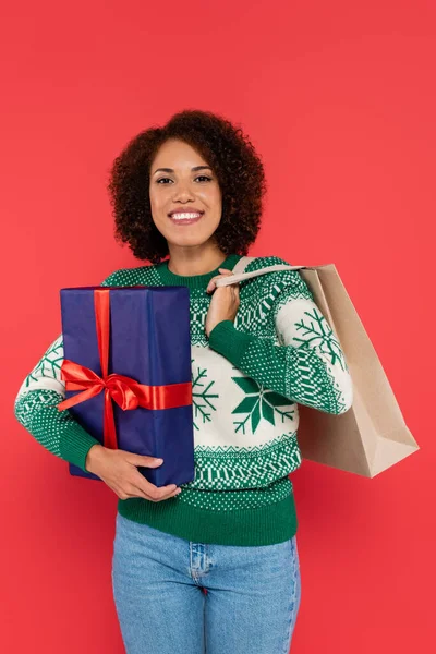 幸せなアフリカ系アメリカ人女性で暖かいセーターを保持ショッピングバッグと青ギフトボックス孤立した上赤 — ストック写真