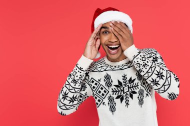 amazed african american man in santa hat and winter sweater hat peeping isolated on red clipart