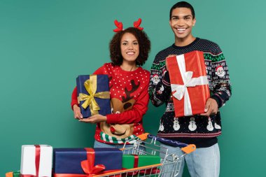 Mutlu Afro-Amerikan çifti ellerinde Noel hediyeleriyle alışveriş arabasının yanında.