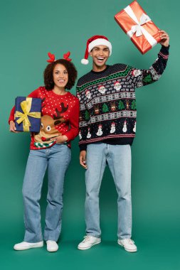 Heyecanlı Afro-Amerikan çift Noel Baba şapkası ve ren geyiği boynuzları yeşil arka planda hediye kutuları tutuyorlar.