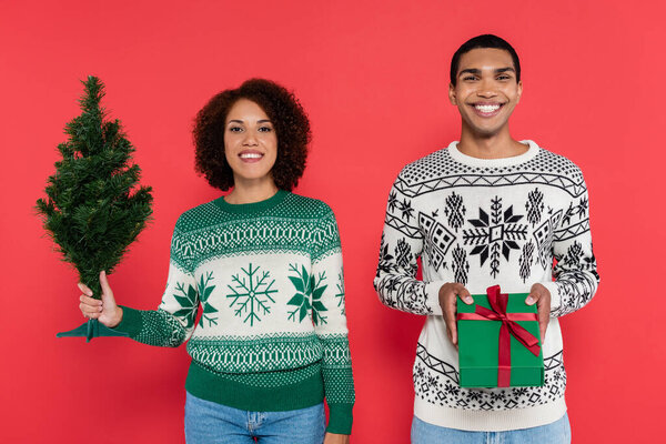 young and stylish african american couple with gift box and small christmas tree looking at camera isolated on red