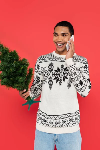 joyful african american man in sweater with winter ornament holding small christmas tree while talking on cellphone isolated on red