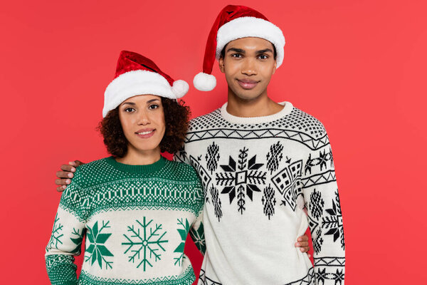 smiling african american couple in santa hats and sweaters with winter ornament isolated on red