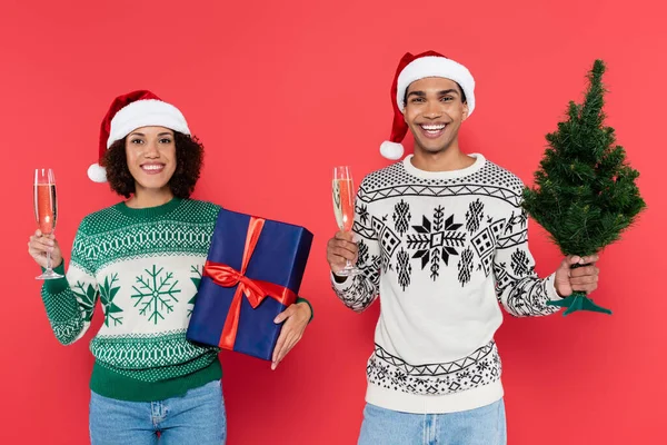 陽気なアフリカ系アメリカ人のカップルがシャンパングラスを持ちクリスマスツリーとブルーギフトボックスを赤で隔てて — ストック写真