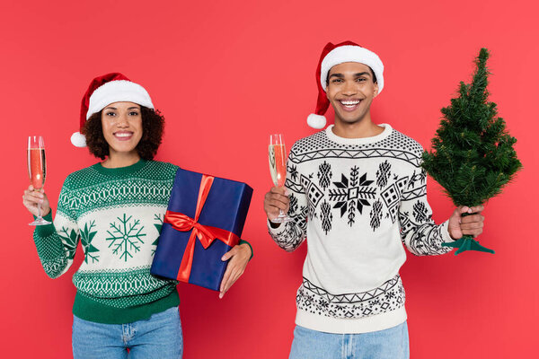 cheerful african american couple holding champagne glasses with christmas tree and blue gift box isolated on red