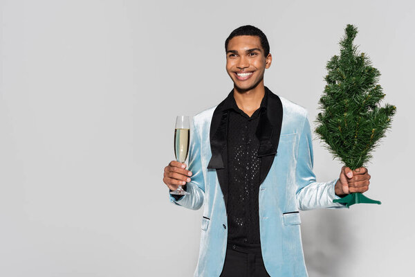 elegant african american man with champagne glass and small christmas tree smiling and looking away on grey background