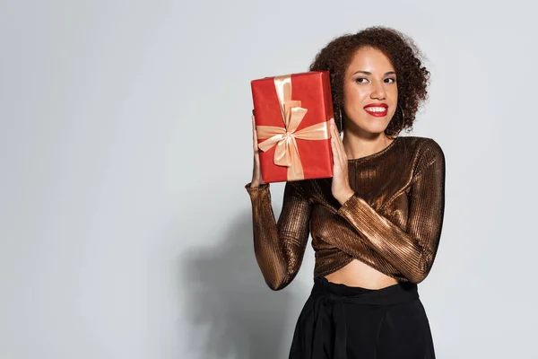 幸せなアフリカ系アメリカ人女性で光沢のあるブラウス保持赤ギフトボックス上の灰色の背景 — ストック写真