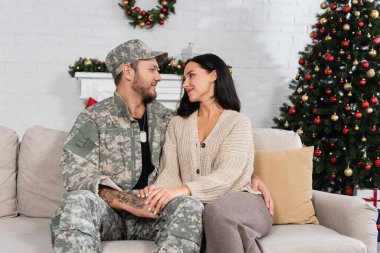 Kamuflajlı adam, süslü Noel ağacının yanındaki koltukta otururken mutlu karısını kucaklıyor.
