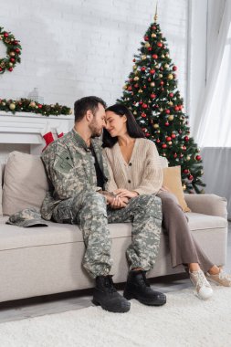 Kamuflajlı adam mutlu eşiyle el ele tutuşurken aynı zamanda süslü Noel ağacının yanındaki kanepede yüz yüze oturuyor.