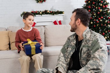Mutlu çocuk, babasının yanında Noel hediyesiyle koltukta oturuyor. Oturma odasında kamuflajlı, şenlikli bir dekorasyonla.