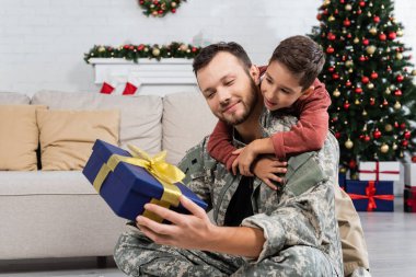Kamuflajlı babayı kucaklayan çocuk oturma odasında elinde Noel dekoruyla hediye kutusuyla bekliyor.