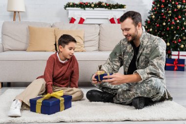 Oğlunun yanında oturan gülümseyen bir asker ve oturma odasında Noel süsleriyle hediye kutuları.