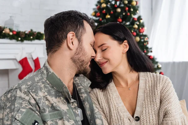 brunette woman with closed eyes smiling near husband in military uniform near blurred christmas tree