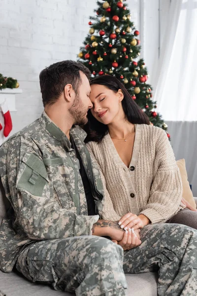 military man and smiling woman with closed eyes sitting face to face near blurred christmas tree at home