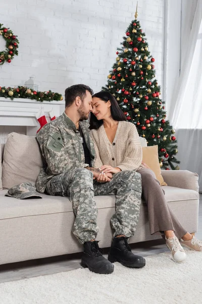 man in camouflage holding hands with happy wife while sitting face to face on couch near decorated christmas tree