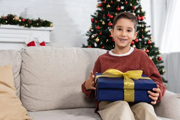 陽気な男の子とクリスマスプレゼントソファに座ってお祝いの装飾が施されたリビングルームでカメラを見て — ストック写真