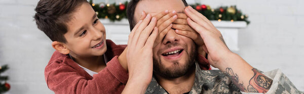 cheerful boy playing guess who game with smiling dad in camouflage and covering his eyes with hands, banner