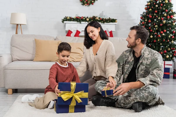 happy boy opening gift box while sitting on floor near smiling mother and father in military uniform