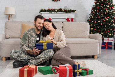 Kocası üniformalı mutlu bir kadın hediye kutularının yanında oturuyor ve Noel ağacı süslüyor.